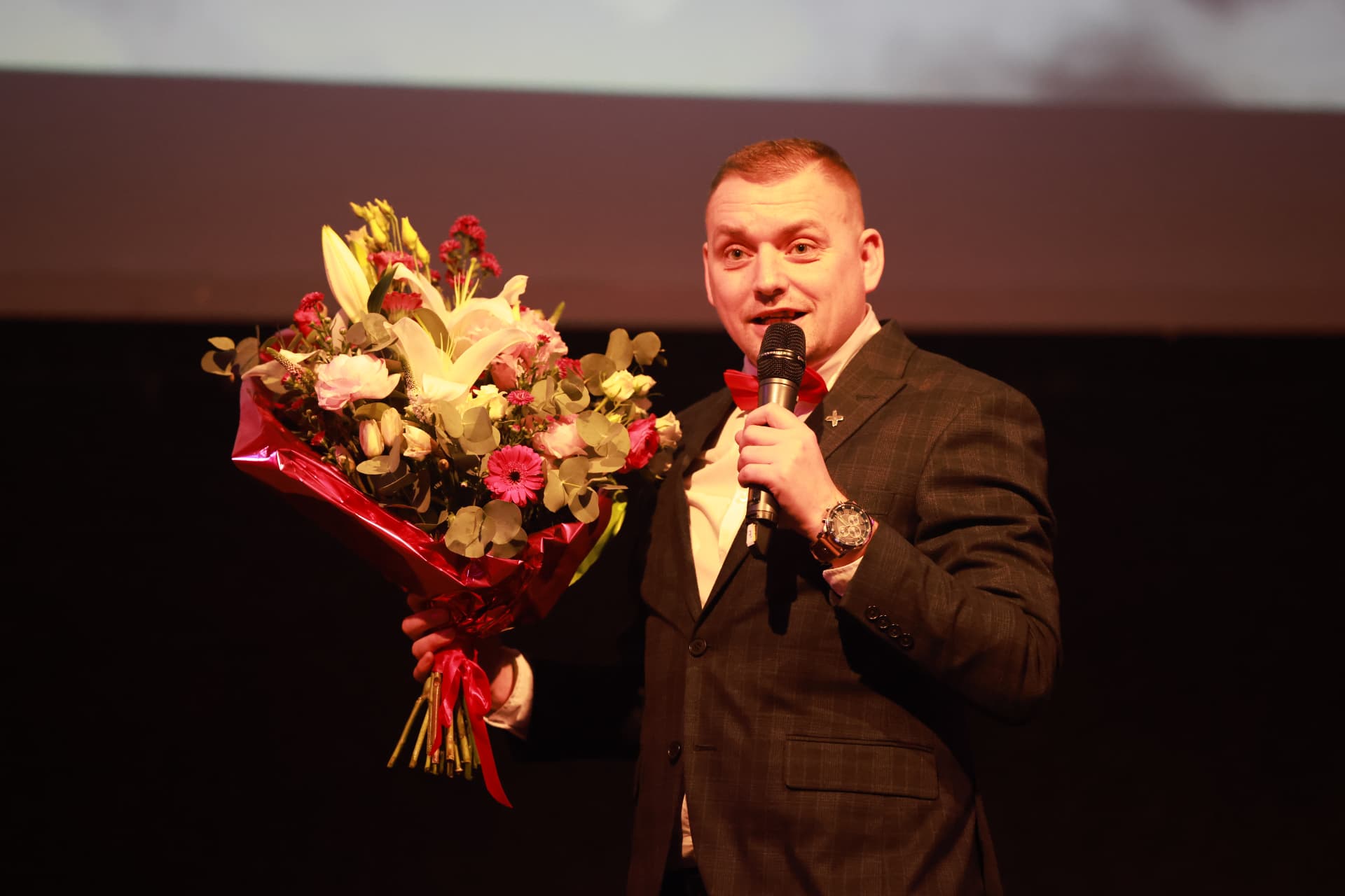 Moderátor s kyticí a mikrofonem na pódiu 2. charitativního plesu